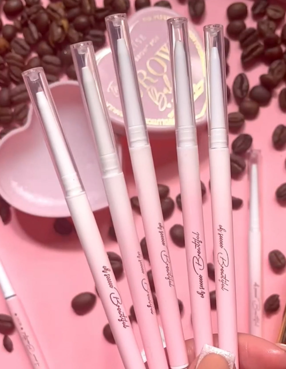 A collection of pink P.Louise eyebrow pencils with spoolie brushes, displayed alongside coffee beans and a pink dish.