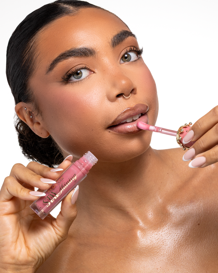 A close-up of a woman applying a pink P.Louise Stay Shining Lip Oil liquid lip product with a doe-foot applicator while holding the tube in her other hand, set against a clean white background.