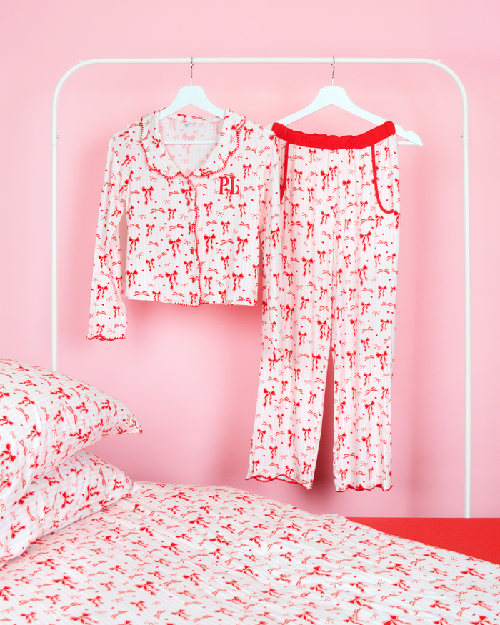 Set de pantaloni de pijama cu fundă de Crăciun P.Louise pentru fete, cu un design cu fundă roșie peste dungi roz și albe, atârnați pe umerase într-un dormitor roz cu lenjerie de pat asortată