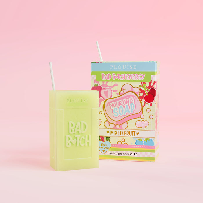 Un săpun verde cu „Bad Bitch” gravat pe ea, însoțit de un ambalaj colorat cu sucuri cu eticheta „Mixed Fruit”, prezentat pe un fundal roz.