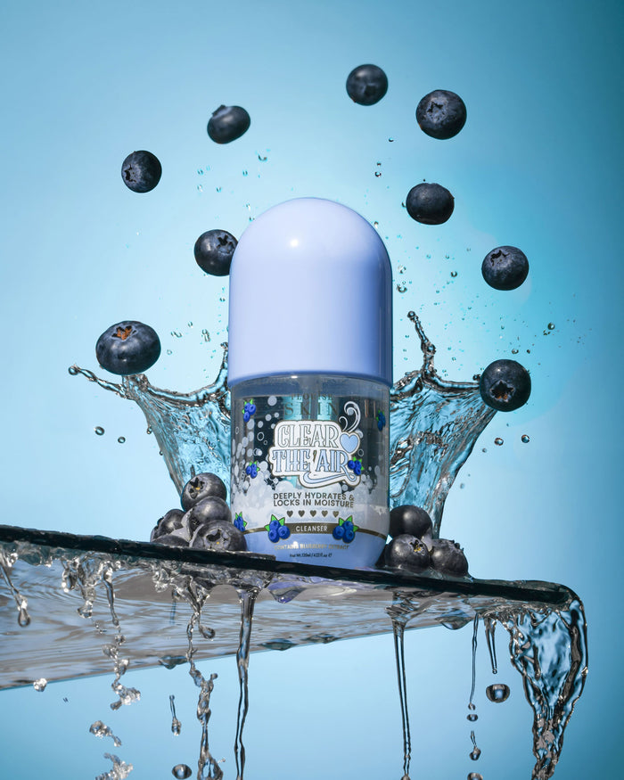Sticla de P. Louise Clear The Air Blueberry Gel Cleanserl înconjurată de afine și stropi de apă pe un fundal albastru